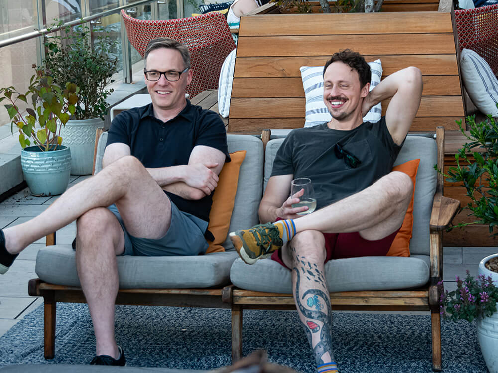 Two employees sitting with their legs crossed next to one another smiling in conversation on a patio couch.