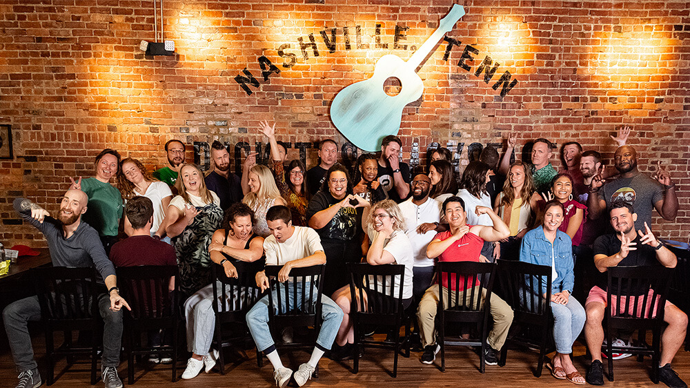 A company team photo of 30+ people doing a variety of silly faces and poses in front of brick wall that reads 