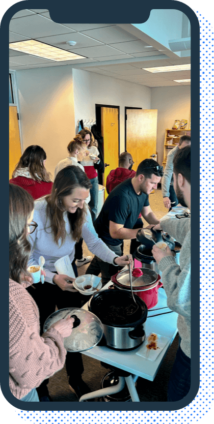 A phone with with a photo of a group of people in an office setting serving themselves chili from a table lined with crockpots.